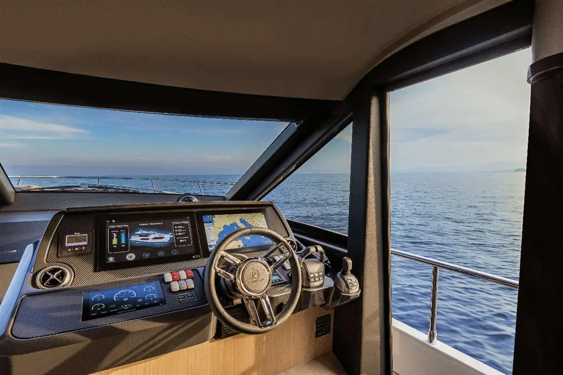 Interior of a modern yacht’s cockpit with a steering wheel, control panels, and a view of calm waters under a clear sky.